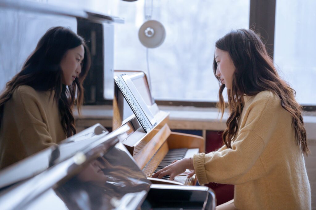 Side view of focused ethnic female musician sitting near piano and pressing keys in daylight from big window