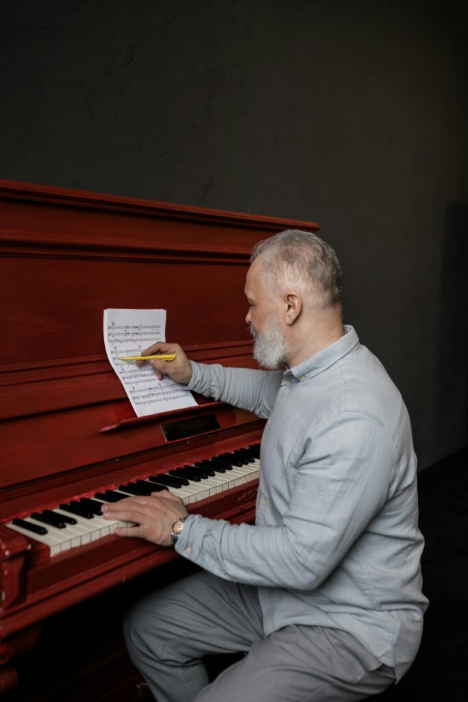 Senior man writing music on a red piano indoors, focusing on musical composition.