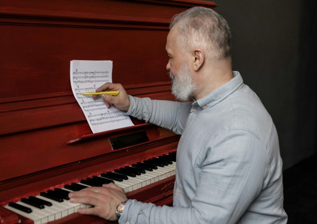 Senior man writing music on a red piano indoors, focusing on musical composition.