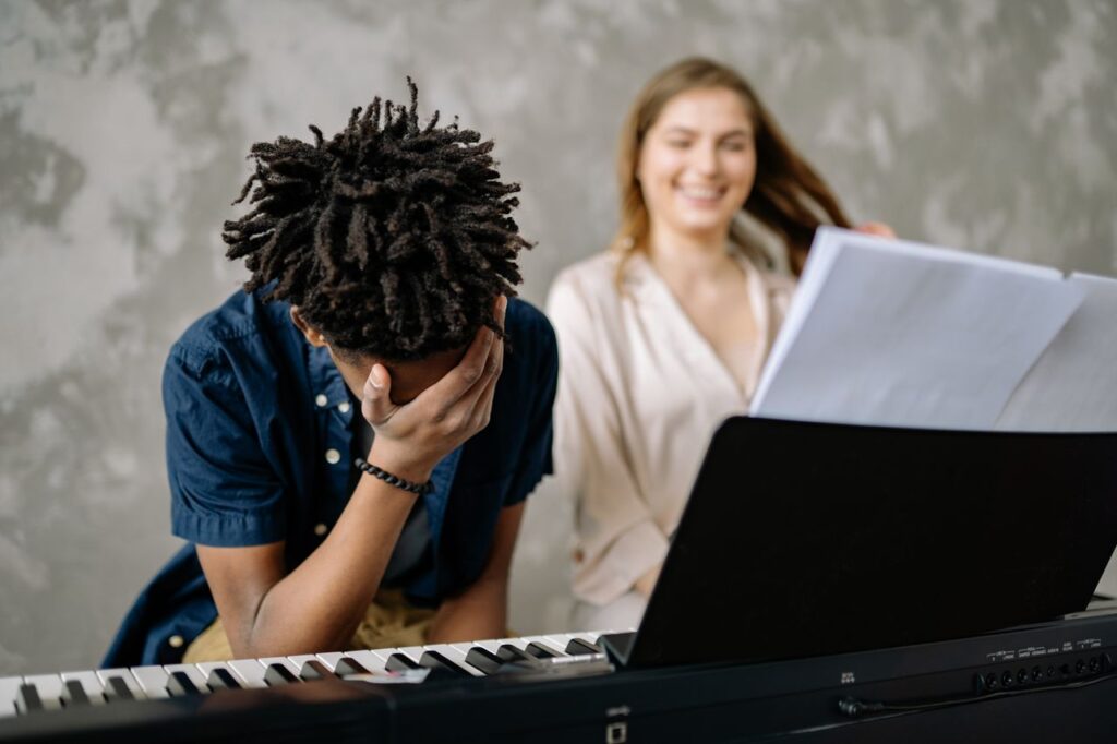 Young boy feeling overwhelmed at piano lesson with supportive teacher nearby.